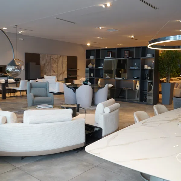 Modern living room showroom with white curved sofas, gray armchairs, marble table, and decorative lighting fixtures.