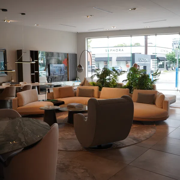 Modern showroom with curved beige sofas, armchairs, glass tables, and large windows overlooking city stores.