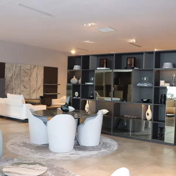 Modern living room with white chairs, round glass table, gray shelving unit, and indoor plants under soft lighting