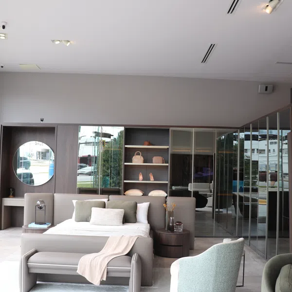 Modern bedroom showroom with neutral tones, bed, armchairs, round mirror, and decorative shelving under bright lighting.