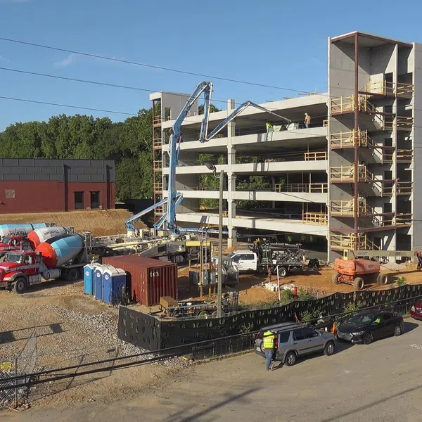 Concrete parking garage under construction with cement trucks, workers, and equipment on site during daytime.