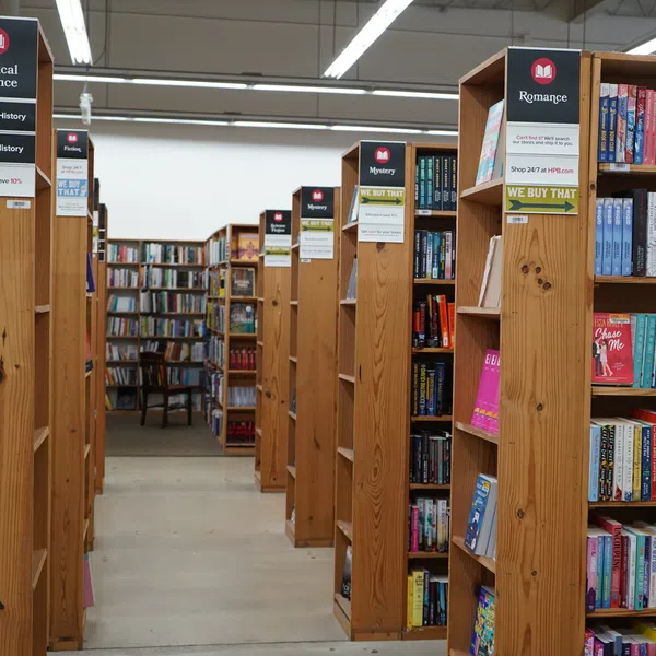 A bookstore aisle with wooden shelves labeled Political Science, Mystery, and Romance filled with books under bright overhead lights.