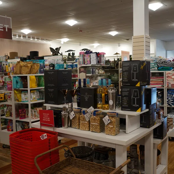Gourmet food and gift items displayed on white shelves with red shopping baskets in a home goods store.