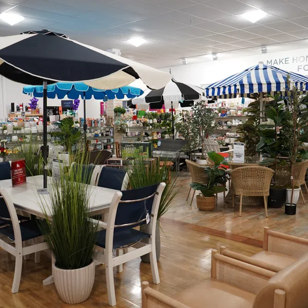 Indoor furniture store display featuring patio tables with umbrellas, chairs, and potted plants on wooden floor.