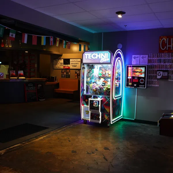 Neon-lit claw machine filled with plush toys in a dim arcade with colorful flags and a Cheap Thrills sign.