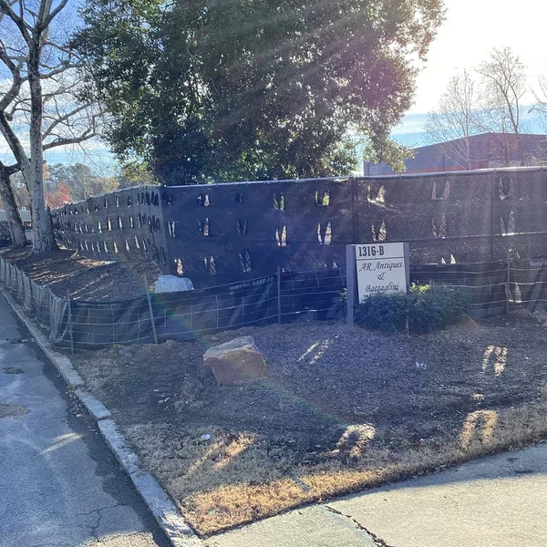 Construction site fenced with black netting along a sidewalk under clear blue sky and leafless trees.