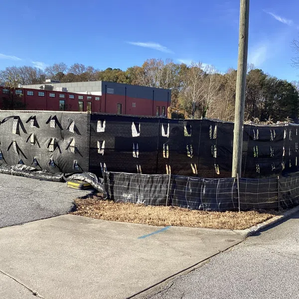 Black construction fence with taped seams surrounding site near a parking lot and building under clear blue sky.