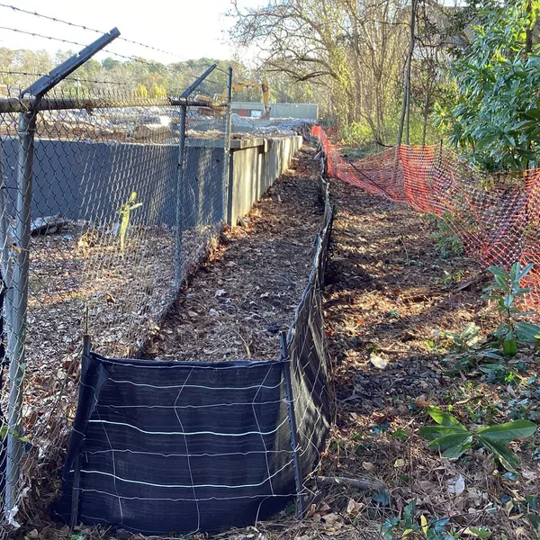 Long narrow dirt path bordered by black silt fencing, chain link fence on left, and orange mesh fencing on right under trees.