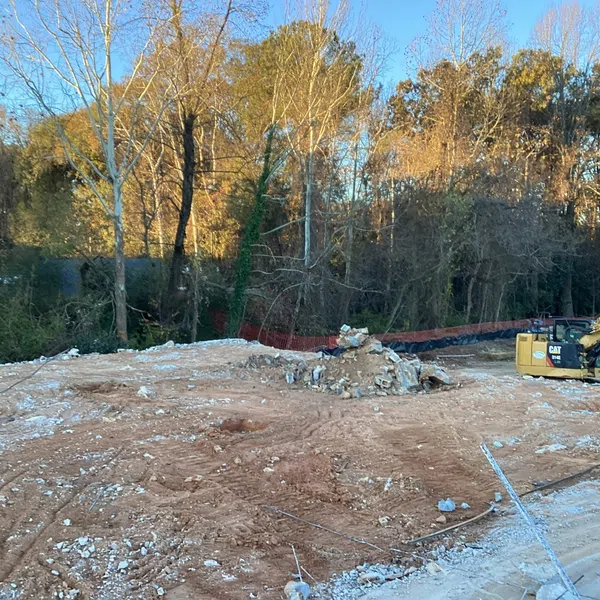 Construction site with excavator, dirt ground, debris piles, and trees in the background during daytime.