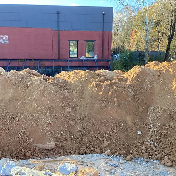 Large dirt mound in front of a brick and gray industrial building with trees in the background