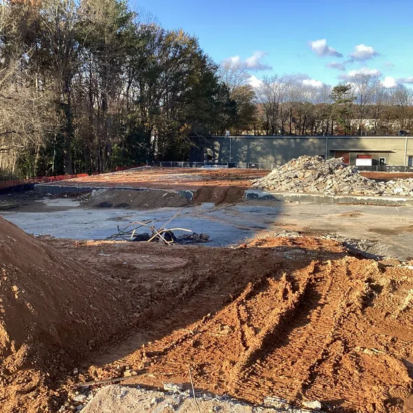 Construction site with earthworks, dirt piles, and debris near a building and trees under blue sky.