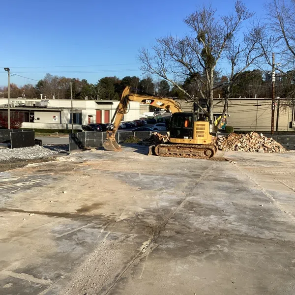 Excavator on cleared construction site with rubble piles and buildings in background under clear sky
