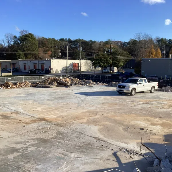 Empty construction site with cleared concrete foundation, debris piles, a white pickup truck, and buildings under clear blue sky.