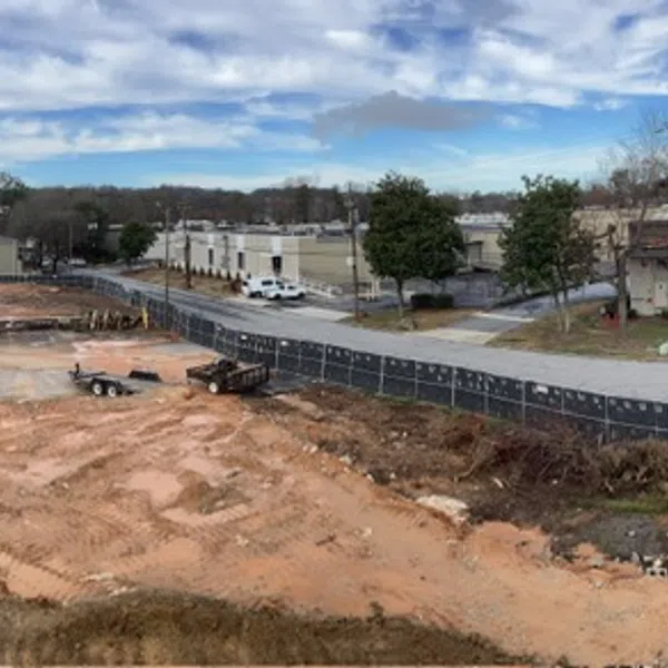 Panoramic view of a construction site with cleared land, equipment, fencing, and adjacent buildings under a partly cloudy sky
