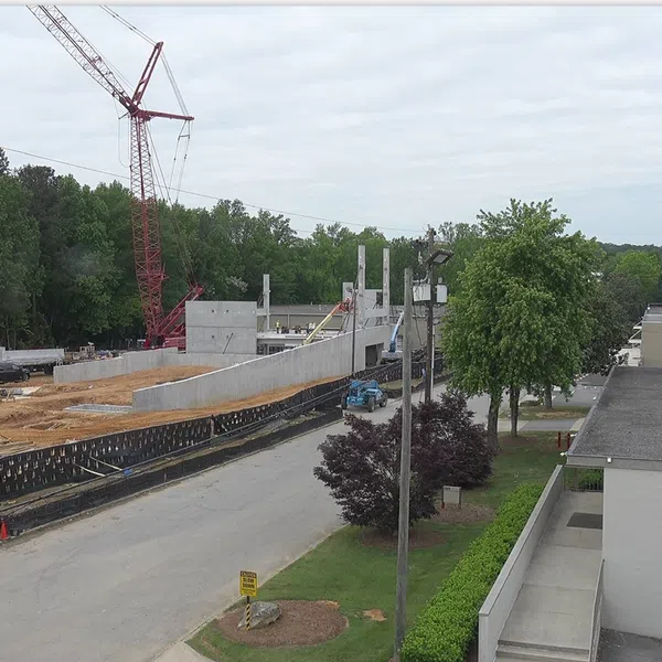 Construction site with crane lifting concrete panels beside road and green trees under a cloudy sky