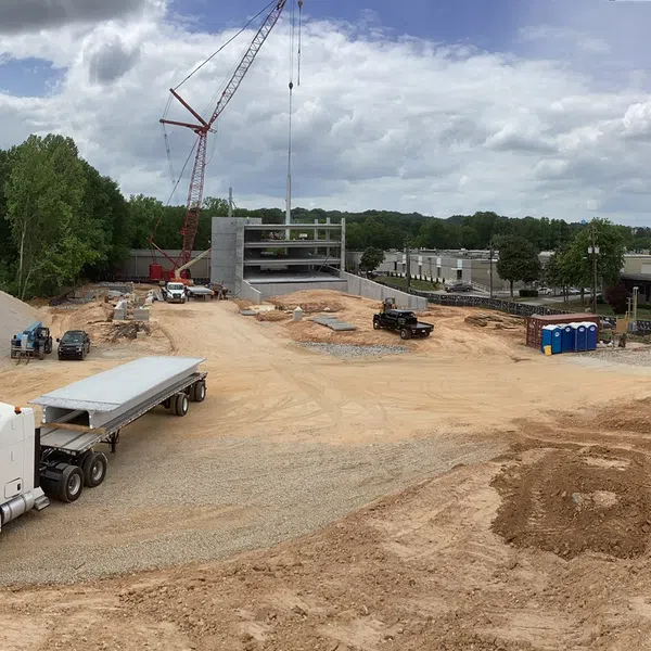 Construction site with crane, flatbed truck, heavy machinery, and building framework under cloudy sky.
