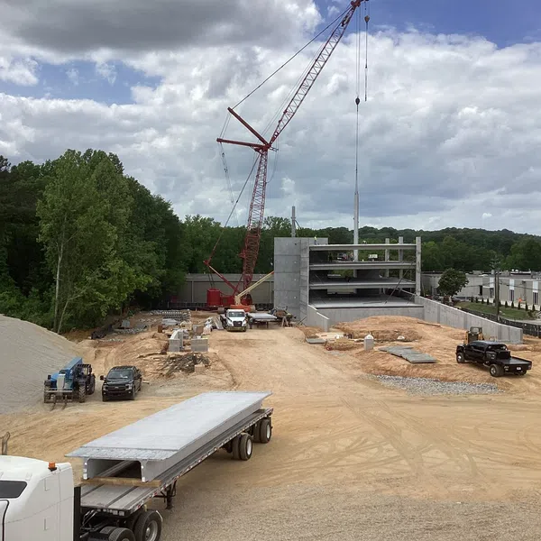 Active construction site with a crane, trucks, and a multi-level concrete parking structure under cloudy skies.
