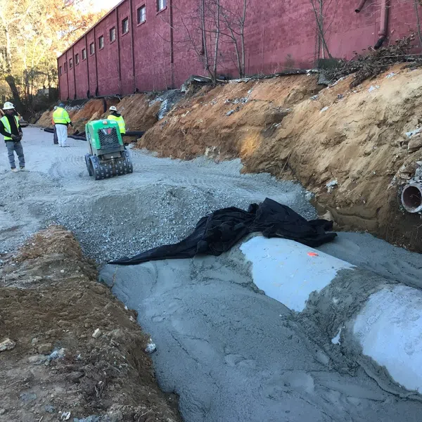Construction workers installing large concrete pipe drainage system along a dirt road near a red brick wall.