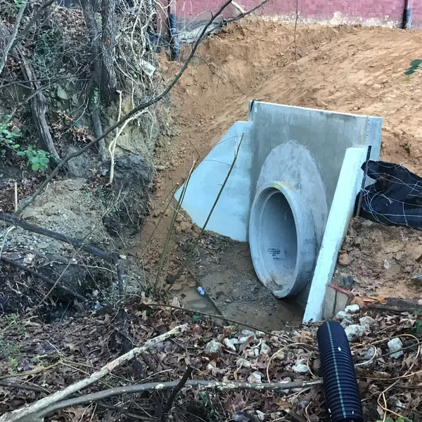 Concrete drainage pipe installed at a forested stormwater runoff site with exposed soil and vegetation.
