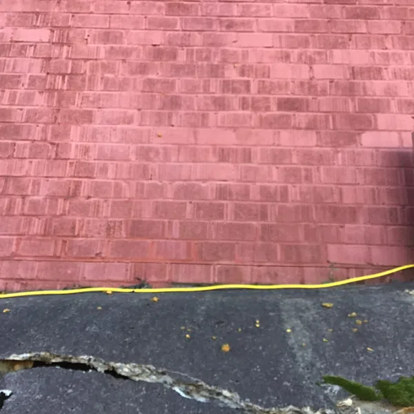 Cracked black asphalt pavement with moss near red brick wall and yellow cable at the base of the wall.