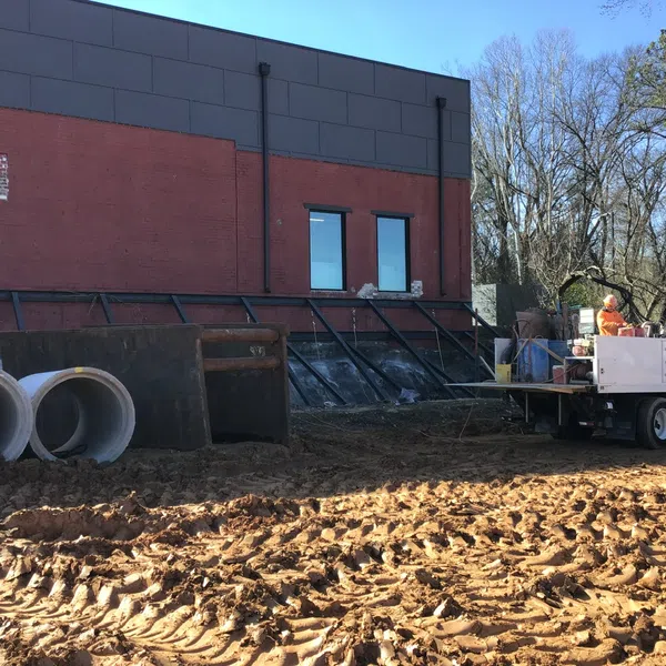 Construction site with large concrete pipes, muddy ground, and a worker operating concrete cutting equipment by a brick building.
