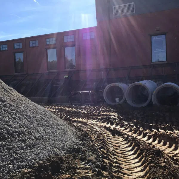 Construction site with gravel pile, large concrete pipes, and tire tracks under bright sunlight near a building.