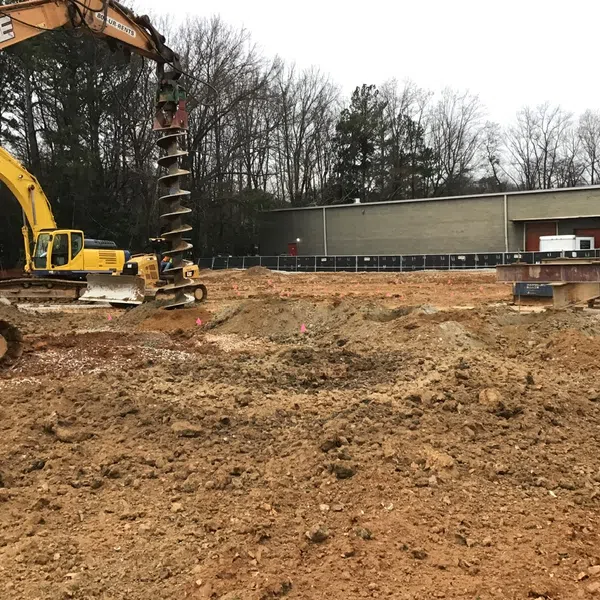Yellow excavator drilling soil on construction site with bare trees and building in background