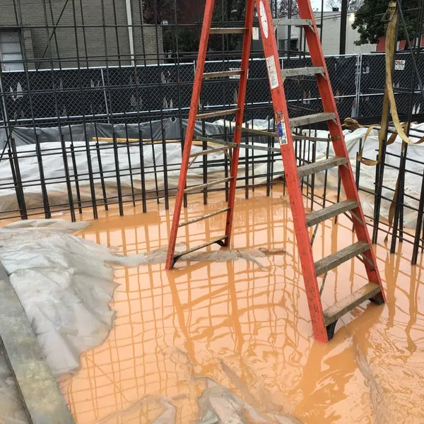 Red ladder stands in muddy water inside a steel rebar framework at a construction site with plastic sheeting nearby