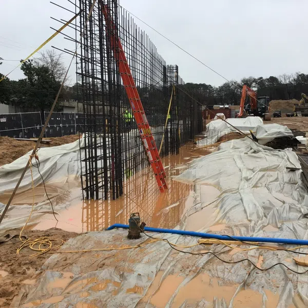 Construction site with steel rebar framework, muddy water, protective plastic sheets, and heavy machinery.