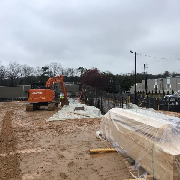 Muddy construction site with an orange excavator, plastic-covered materials, and metal fencing on a cloudy day.