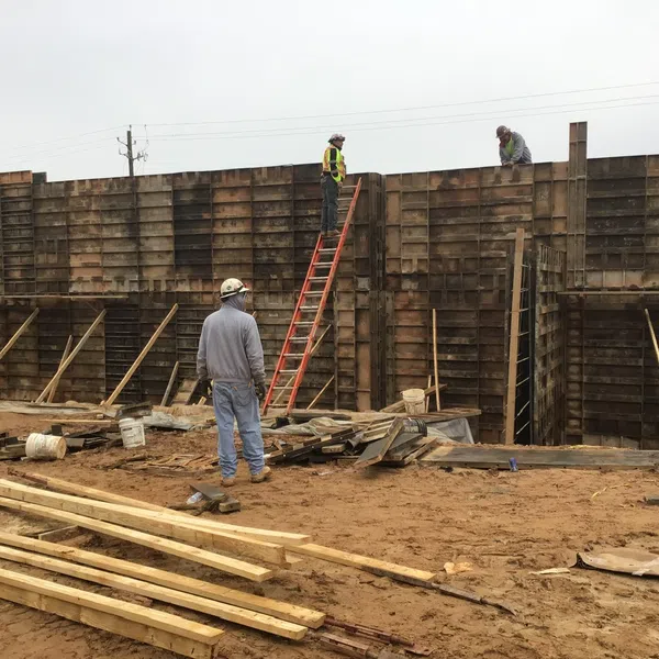 Construction workers building a tall wooden concrete formwork wall on a muddy site under overcast sky.