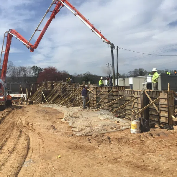 Construction workers pour concrete using a truck-mounted boom pump on a large site with wooden formwork under a blue sky.