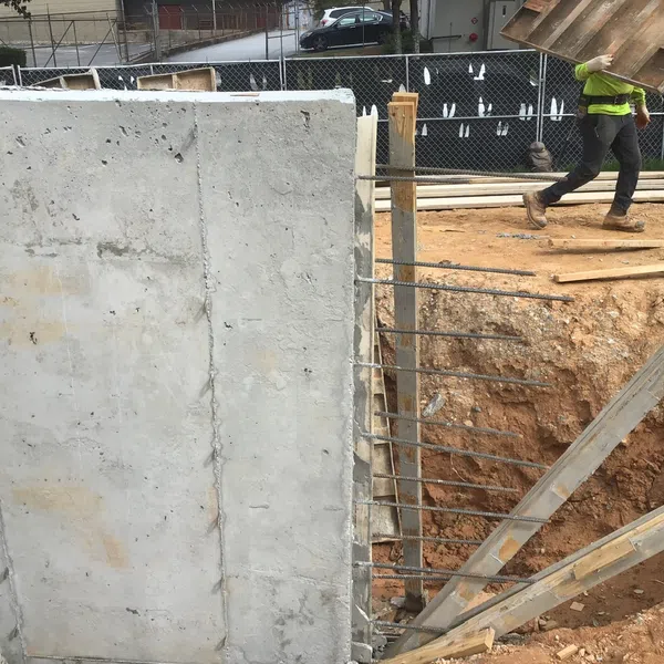 Construction site showing concrete foundation wall with exposed rebar and a worker carrying wooden formwork.