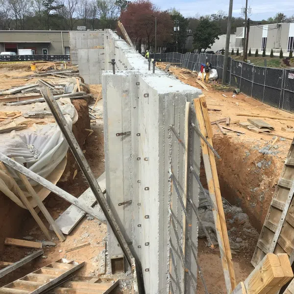 Construction site with concrete retaining walls, wooden supports, and workers preparing excavation area.