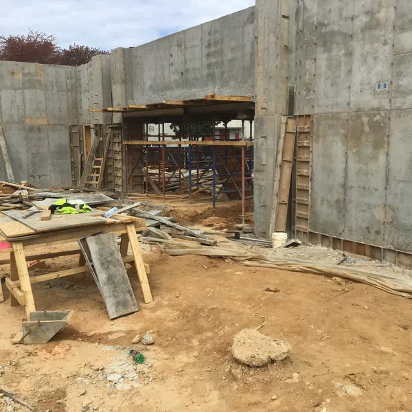 Construction site with concrete walls, wooden scaffolding, and tools scattered on dirt ground under cloudy sky