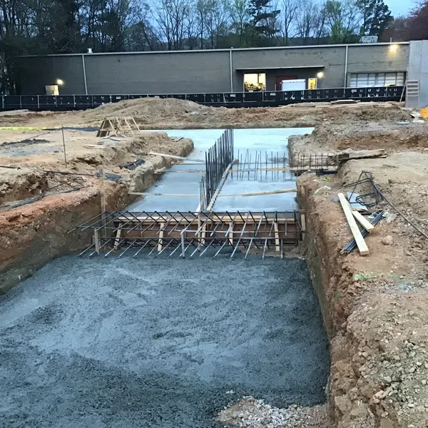 Construction site with concrete foundation and rebar framework in trenches near an industrial building.