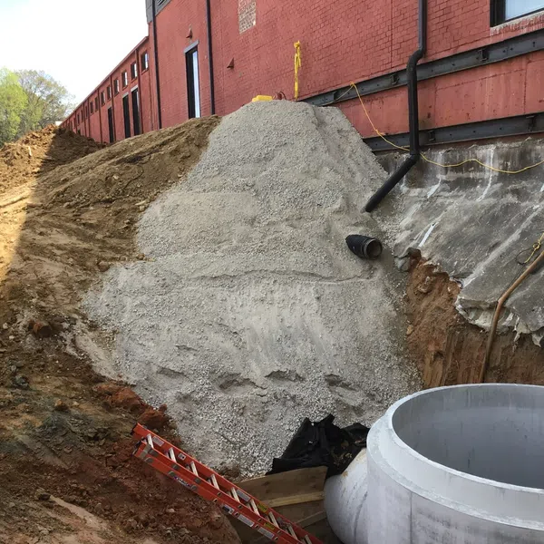 Construction site beside red brick building with gravel mound, drainage pipes, ladder, and concrete pipe section.