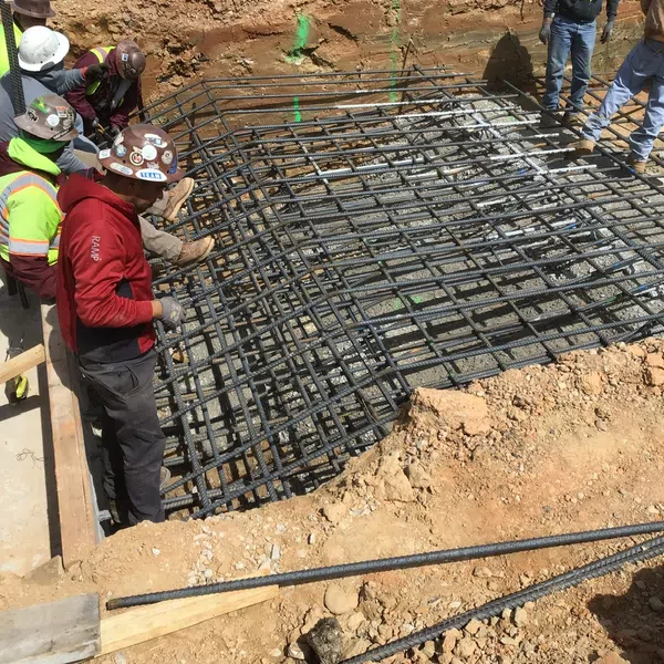 Construction workers install steel rebar grid for concrete foundation in a deep excavation site on a sunny day.