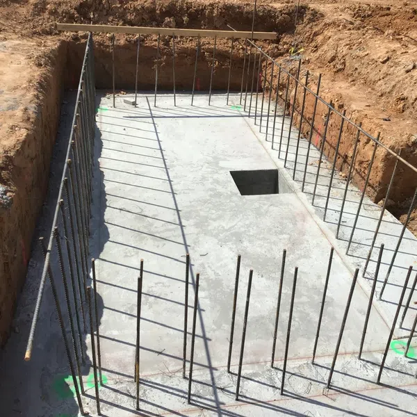 Concrete foundation slab with steel rebar rods protruding, set within an excavated construction site trench.