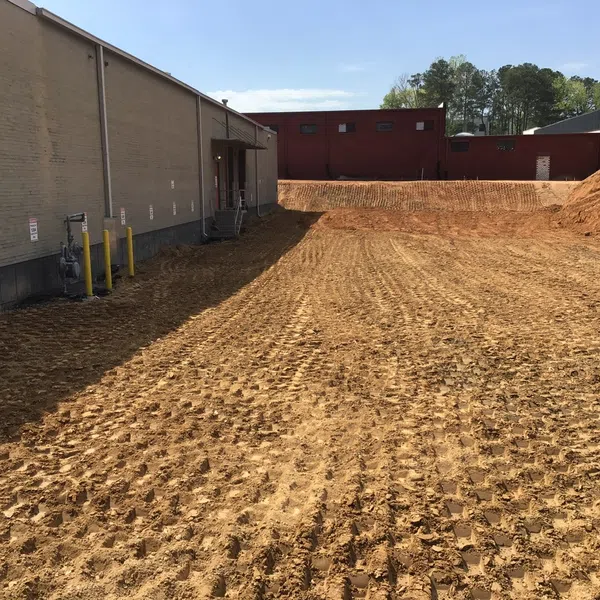 Cleared and graded construction site next to brick buildings with tire tracks on dirt under blue sky.