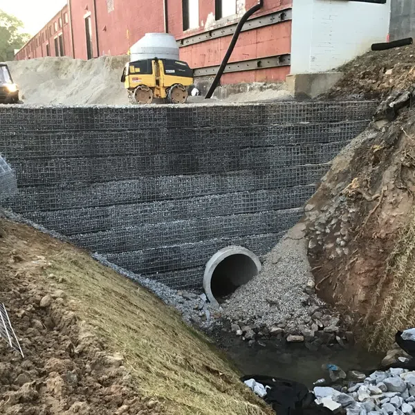 Construction site with concrete drainage pipe and reinforced retaining wall beside sloped earth near building.