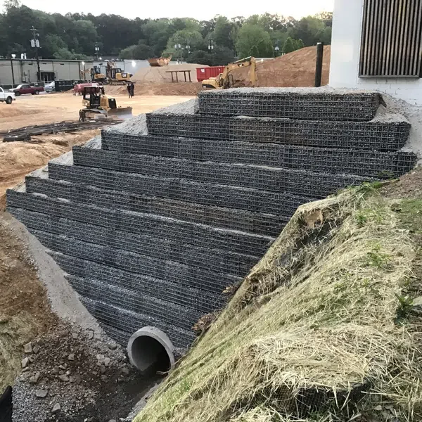 Stepped retaining wall made of wire mesh filled with rocks beside a construction site with machinery and vehicles.