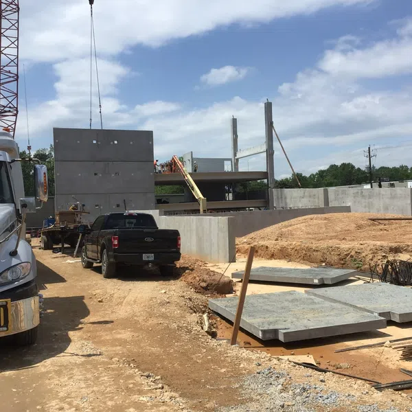 Construction site with trucks, crane lifting concrete panels, dirt road, and building framework under blue sky.
