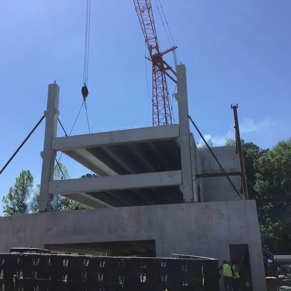 Concrete building structure under construction with crane lifting a floor slab on a clear day with trees nearby