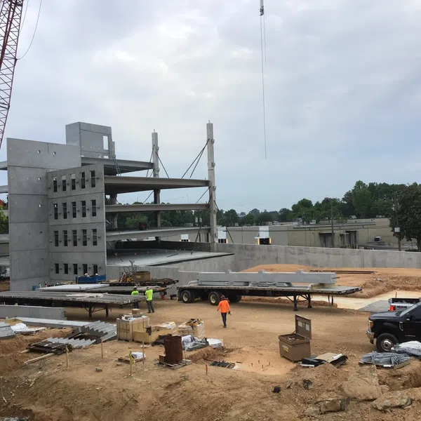 Construction site with concrete building framework, cranes, workers, and construction materials on dirt ground under cloudy sky