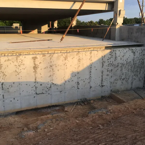 Construction site with concrete foundation walls and exposed rebar under a highway overpass in daylight.