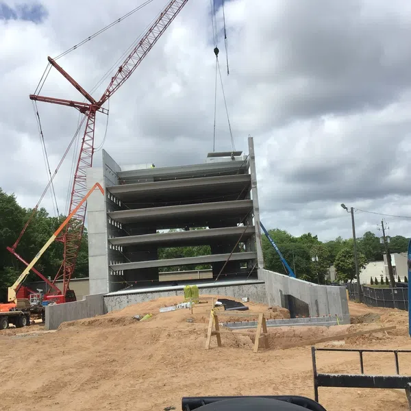 Multi-story concrete parking garage under construction with cranes and dirt foreground on cloudy day