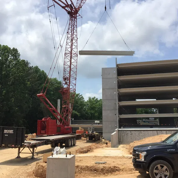 Construction site with crane lifting concrete beam onto multi-story parking structure beside pickup truck under blue sky.