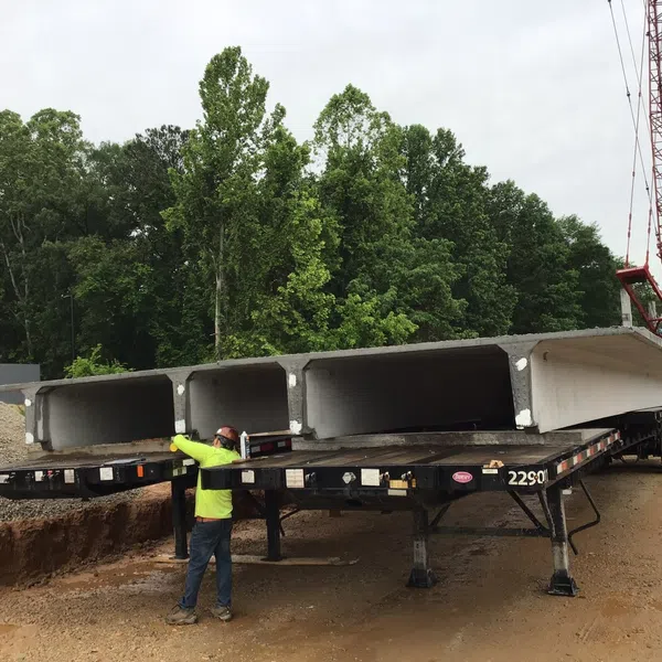 Worker guiding large precast concrete beams on a flatbed trailer at an outdoor construction site with trees in background.