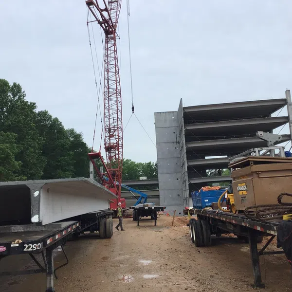 Construction site with crane, flatbed trucks carrying concrete beams, and multi-level concrete parking structure in progress